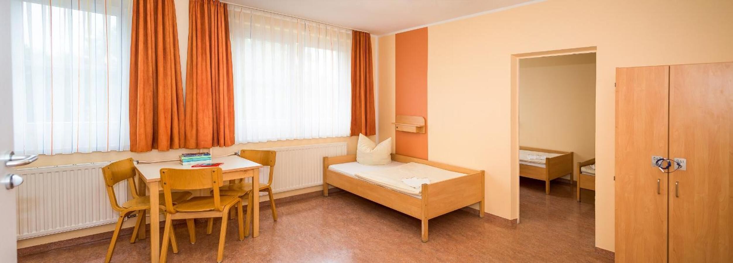 Una habitación pequeña y bien iluminada con una cama de madera, un escritorio con sillas y un radiador.