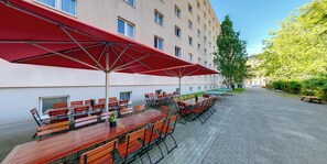 Un espacio al aire libre con mesas y sillas, sombrillas rojas y un edificio al fondo.
