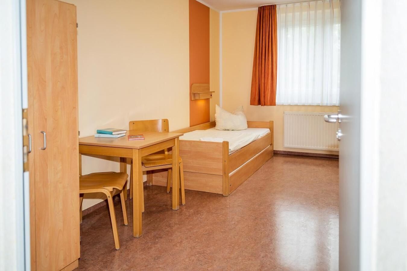 Ein kleiner, gut beleuchteter Raum mit einem Holztisch, einem Einzelbett und einer Heizung.