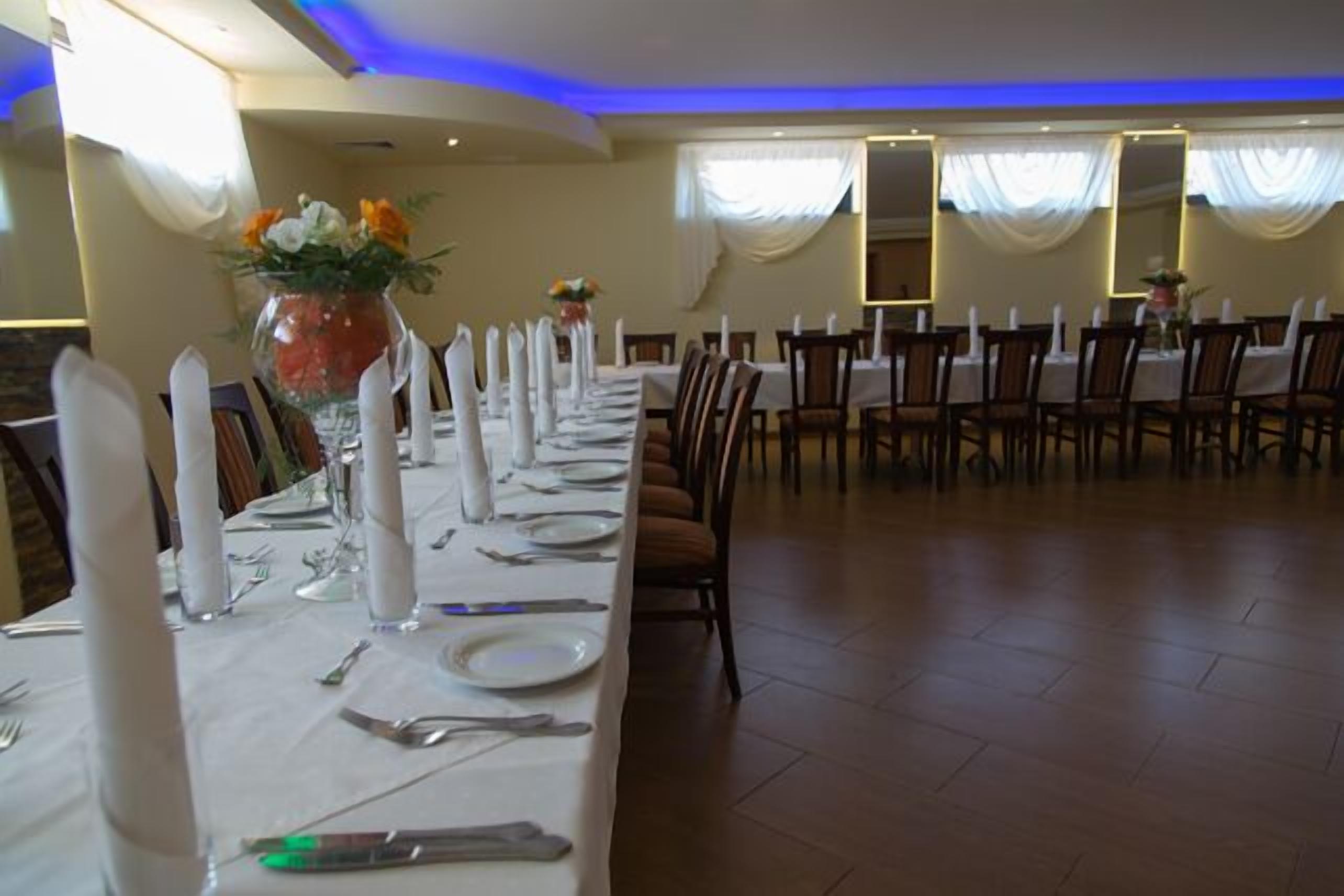 A long dining table set with white tablecloths, silverware, and glassware, flanked by chairs with white napkins.