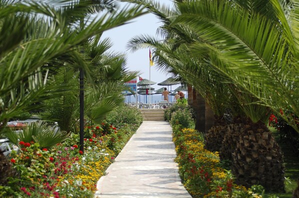 Un sendero bordeado de palmeras y flores de colores que lleva a una zona de playa con sombrillas y sillas reclinables.