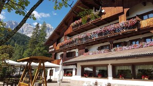 A hotel with multiple balconies adorned with flowers, a patio area with outdoor seating, and a mountainous backdrop. - Hotel Sporting (Val di Zoldo)