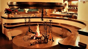 A circular stone fireplace with a fire, surrounded by a curved bench and a hanging light fixture. - Hotel Sporting (Val di Zoldo)