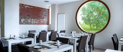A modern dining area with white tables and chairs, a large window with a view of greenery, and a decorative wall with abstract patterns.