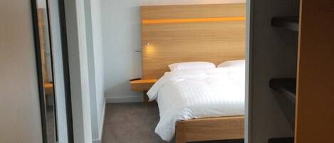 A modern hotel room with a wooden headboard, a bed with white linens, a bedside table, and a built-in shelf unit.