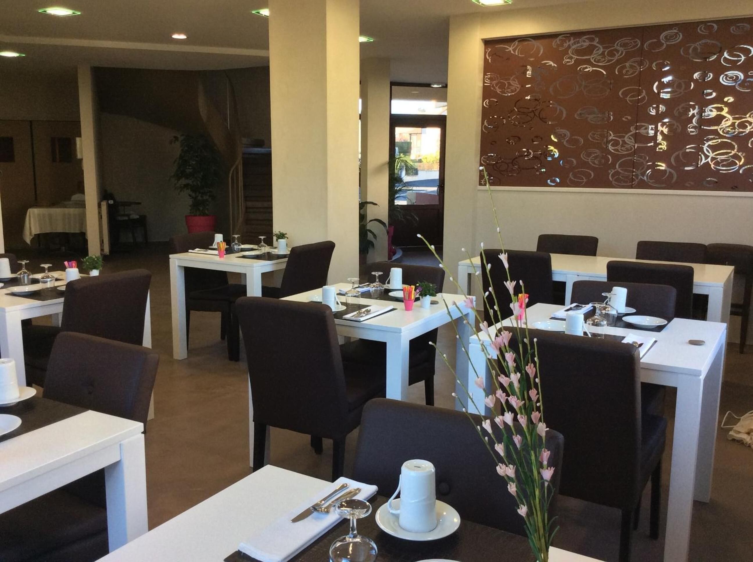 A restaurant with white tables and dark chairs, each set with cutlery and a small vase of flowers.
