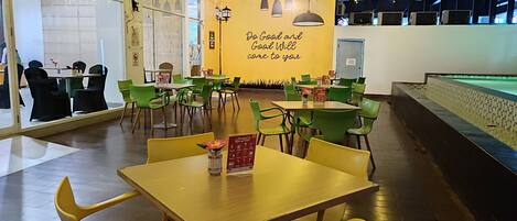 Un restaurante con mesas amarillas y sillas verdes, una pared con una cita inspiradora y luces colgantes en el techo.