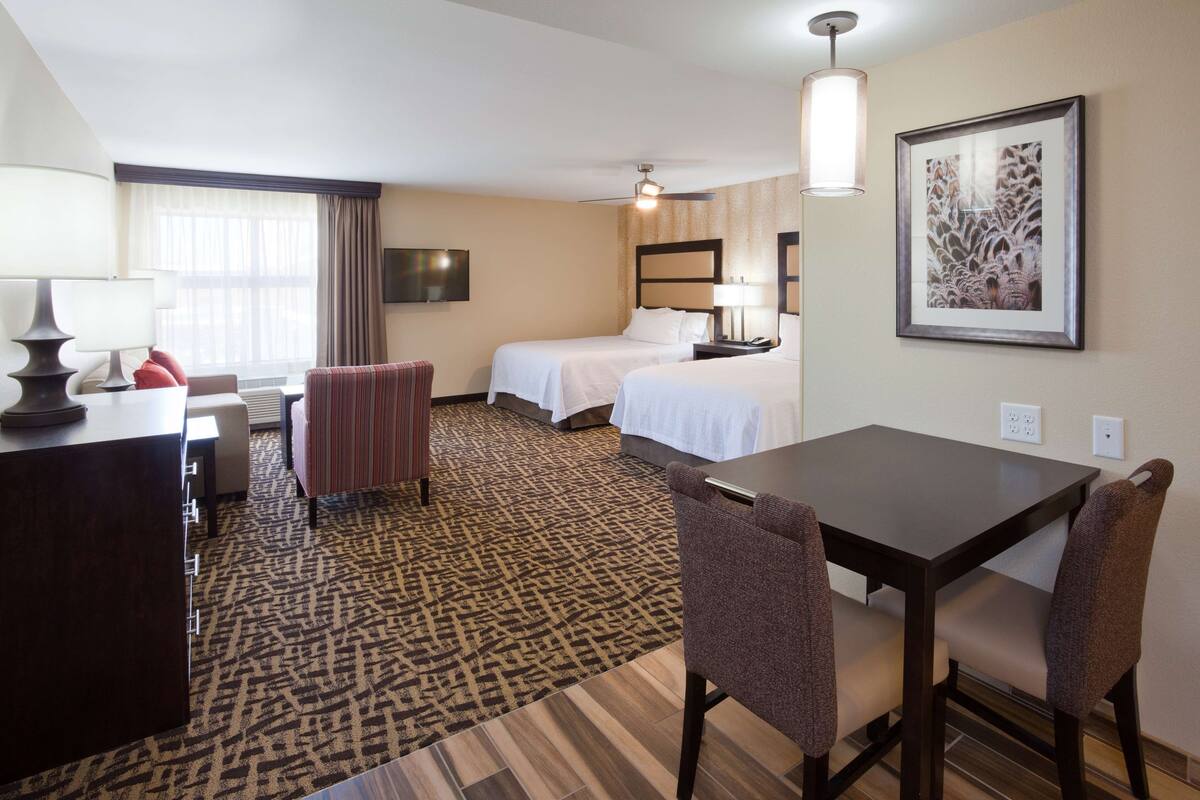 A hotel room with a bed, a desk with a chair, a dining table with chairs, a patterned carpet, and a framed picture on the wall.