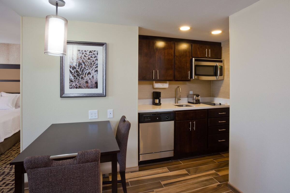 A compact hotel room with a kitchenette, a dining area with a table and chairs, a bed, and a framed artwork on the wall.