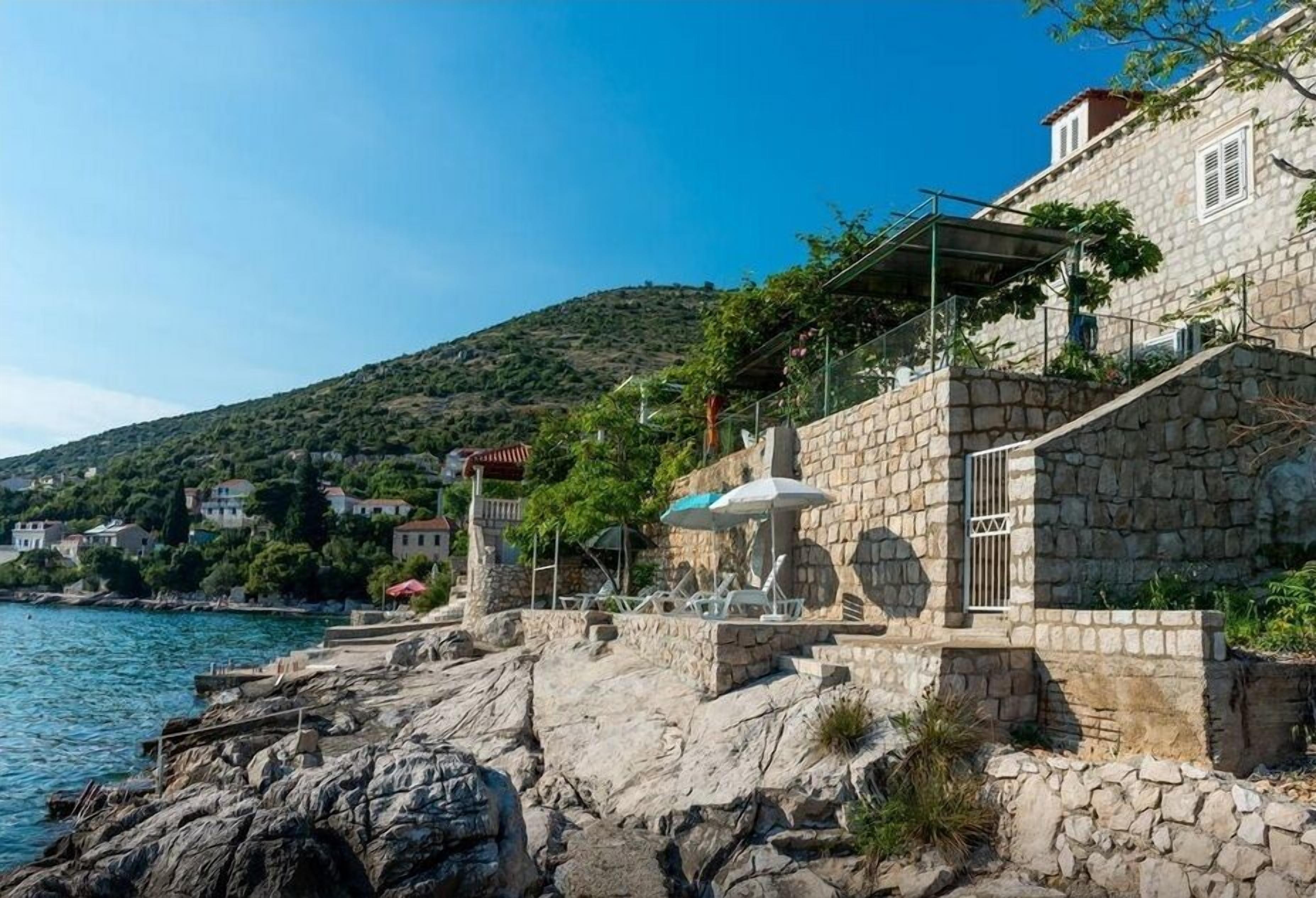 Un edificio de piedra con una terraza que da a una costa rocosa y un mar azul y despejado.