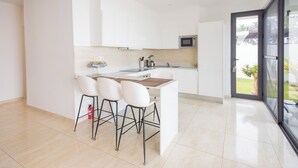 A modern kitchen with white cabinets, a central island with stools, and a view through a glass door to an outdoor area. - Tills Beach Hotel (Gomoa Fetteh)