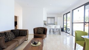 A modern living room with a brown sofa, a coffee table, and a green chair. There is a kitchen area in the background with white cabinets and a dining table. - Tills Beach Hotel (Gomoa Fetteh)