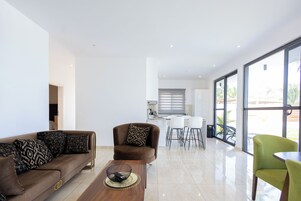 A modern living room with a brown sofa, a coffee table, and a green chair. There is a kitchen area in the background with white cabinets and a dining table.