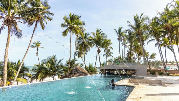 A swimming pool with clear blue water, surrounded by palm trees and a building in the background. - Tills Beach Hotel (Gomoa Fetteh)