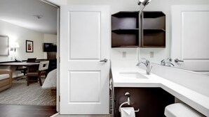 A hotel room with a white door, a desk with a chair, a TV, a bathroom with a sink and mirror, and a shelf above the mirror. - Candlewood Suites Waco by IHG (Waco)