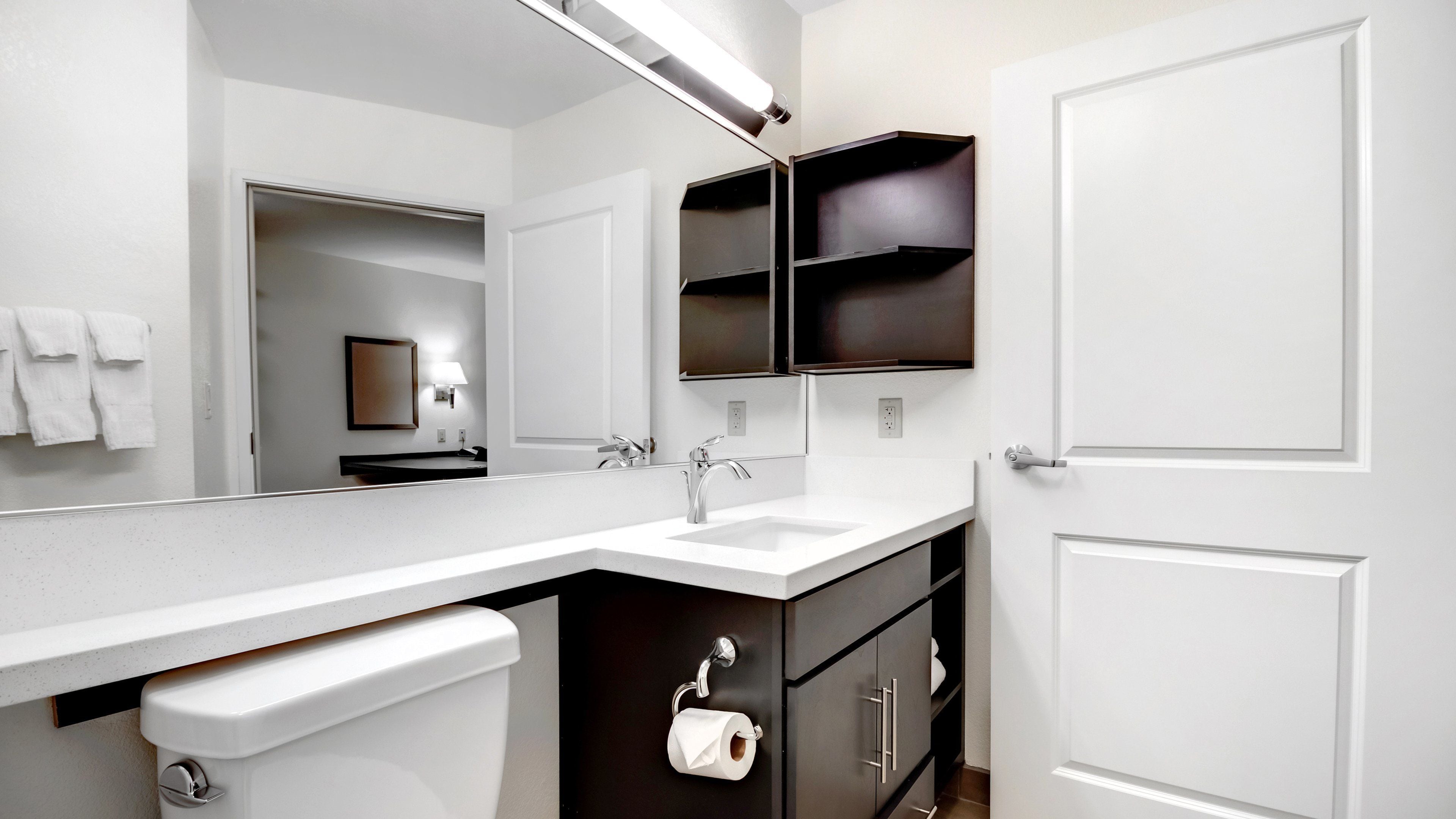 Een moderne badkamer met een witte wastafel, een spiegel en een toilet. Boven de wastafel zijn planken en aan de muur een handdoekhouder.