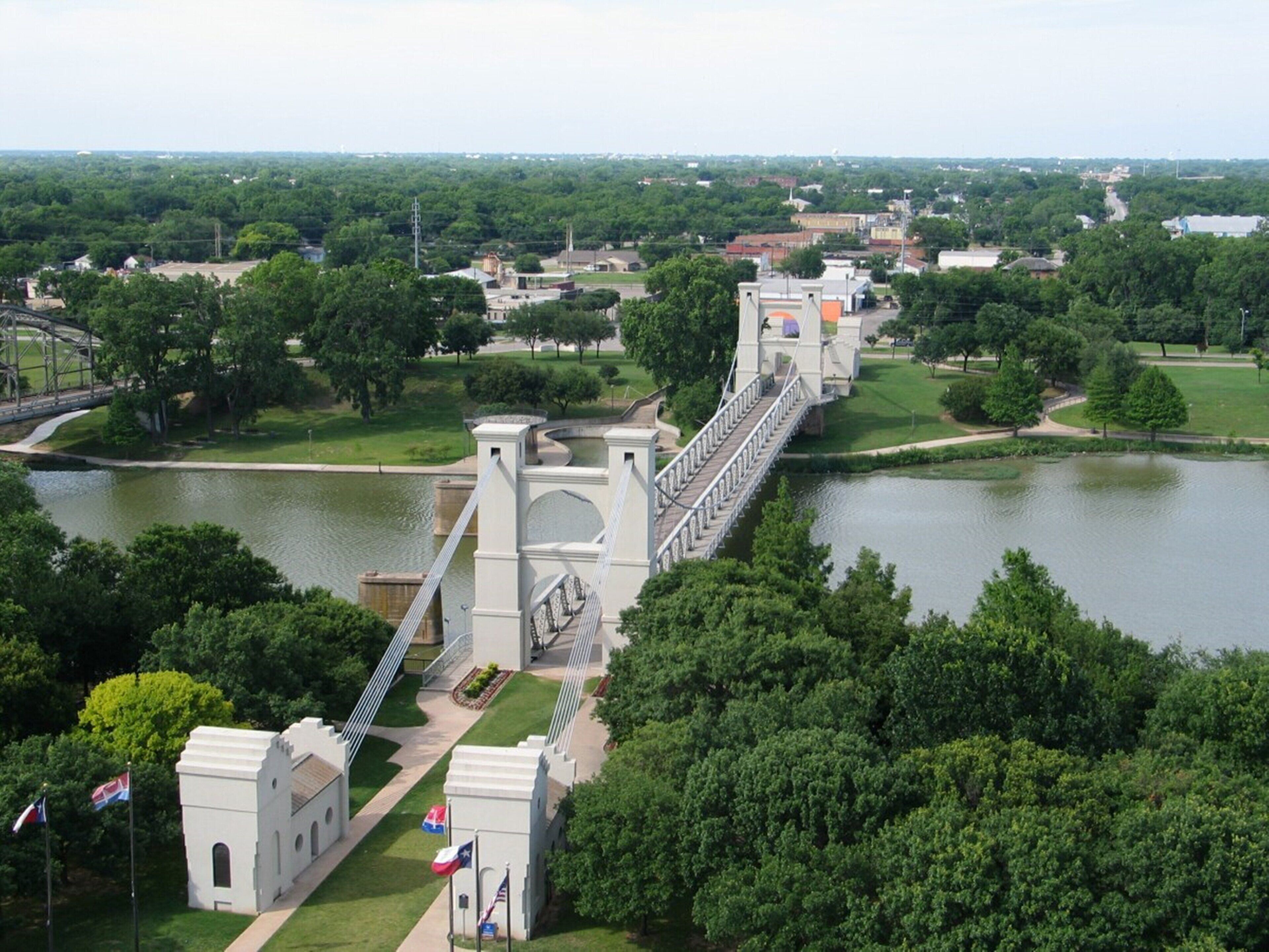 Een grote hangbrug boven een rivier, met een park en gebouwen op de achtergrond.