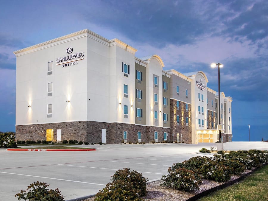 Un hôtel à plusieurs étages, reconnaissable à son grand panneau « Candlewood Suites », doté d’un parking et d’un mât sur lequel flotte le drapeau américain.