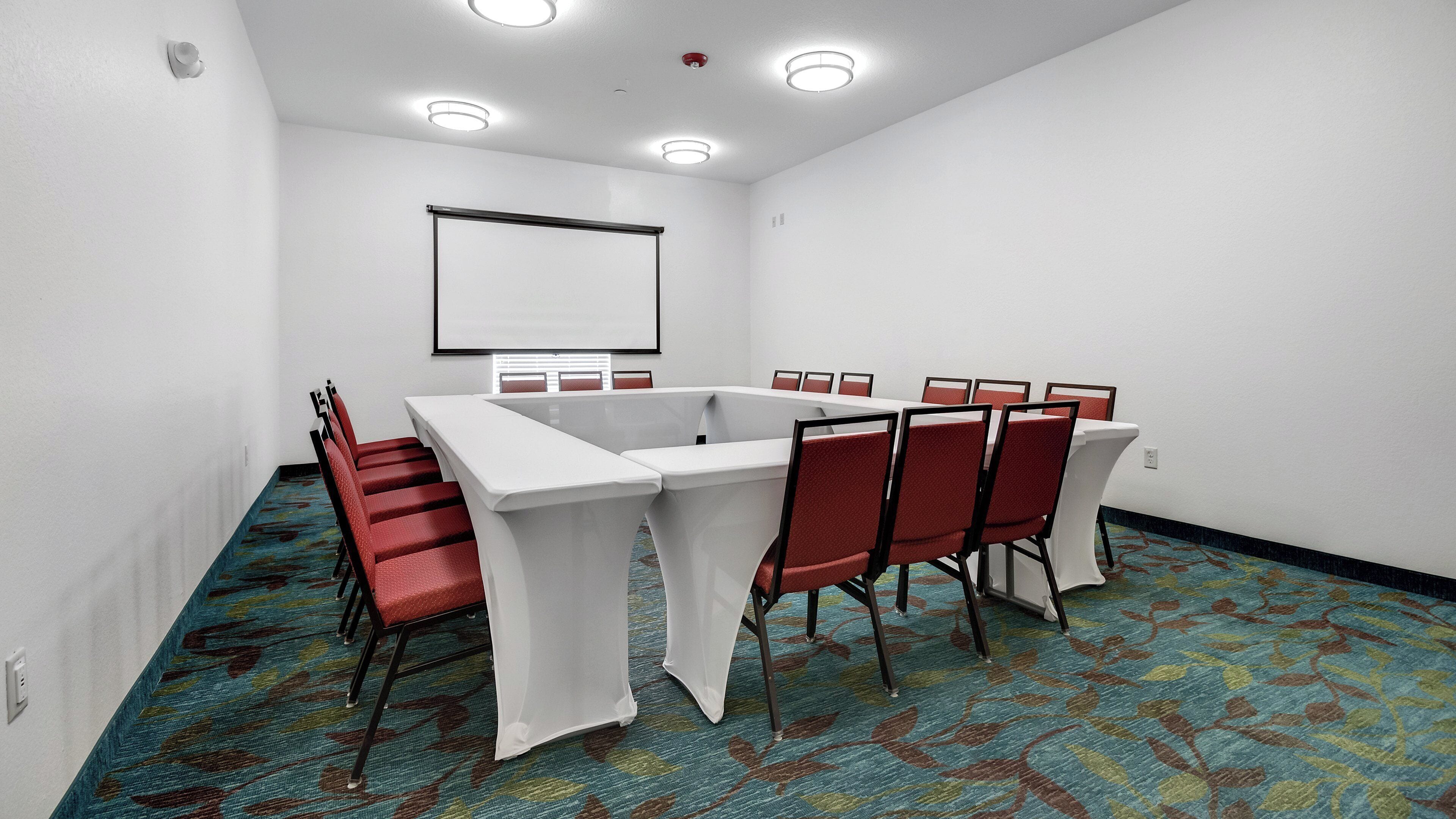Une salle de conférence avec une table blanche, des chaises rouges, un écran de projection et un sol recouvert de moquette.