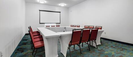 A conference room with a white table, red chairs, a projector screen, and a carpeted floor.