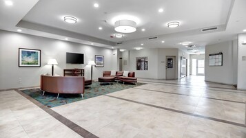 A spacious lobby with a modern design, featuring a seating area with a brown leather sofa and armchairs, a flat-screen TV mounted on the wall, and a patterned rug on the floor.