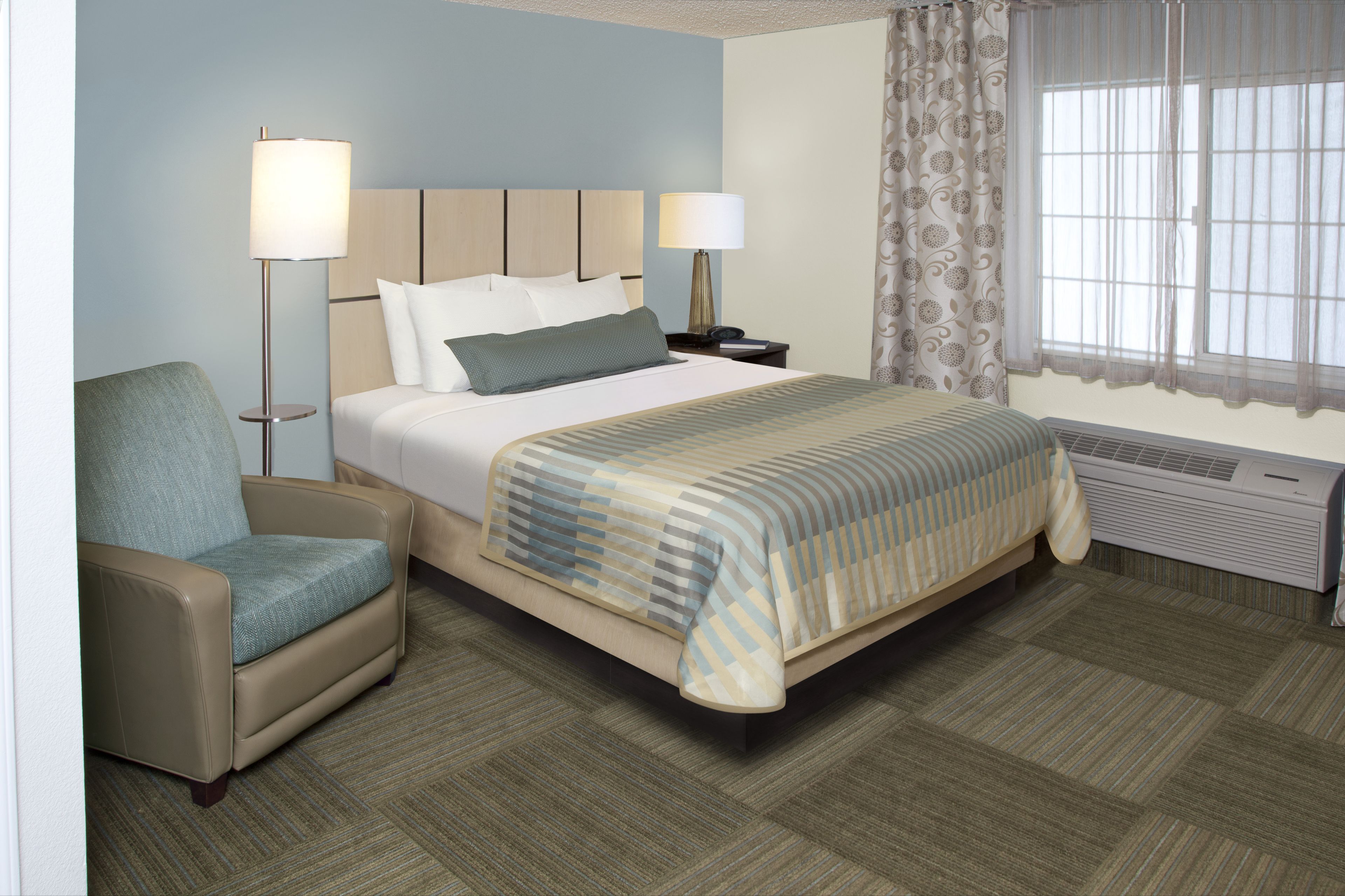 A hotel room with a bed, a chair, two bedside lamps, a window with curtains, and an air conditioning unit.