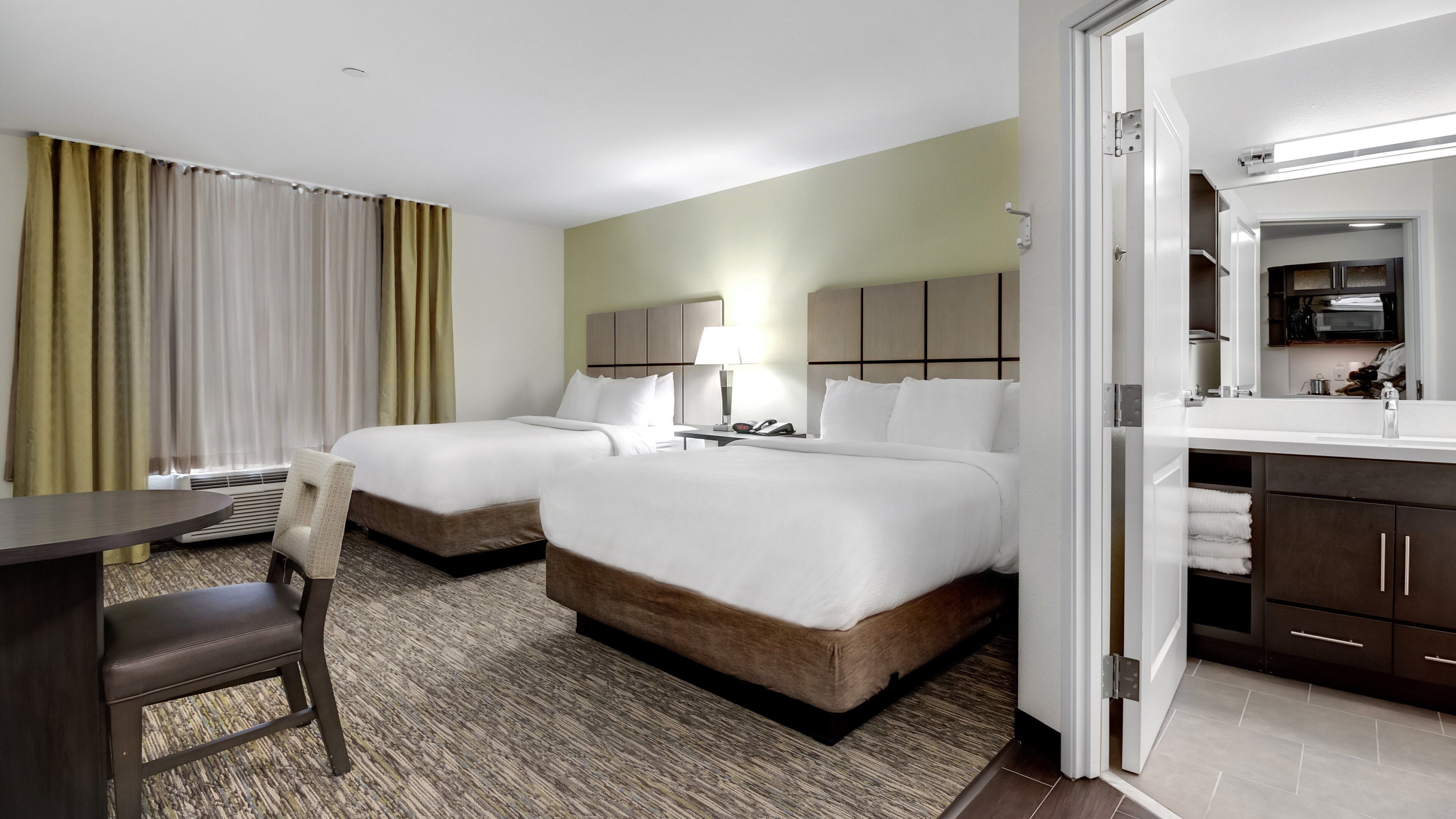 A hotel room with two beds, a desk, a chair, a lamp, a telephone, and a bathroom visible through an open door.