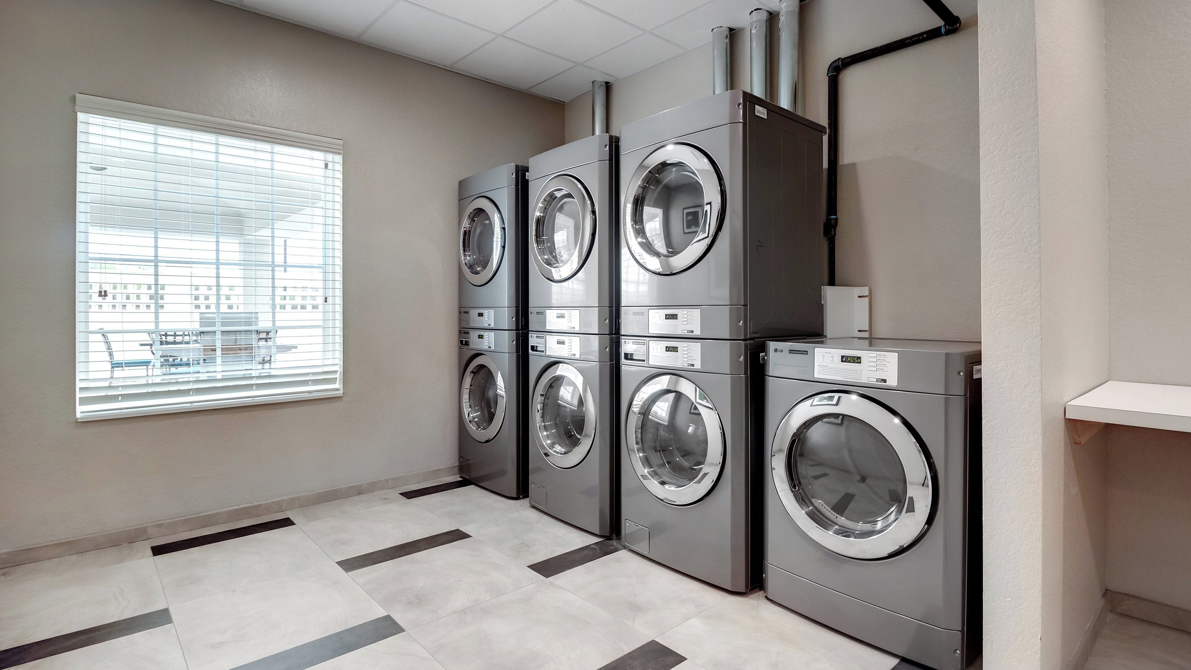 A laundry room with four front-loading washing machines, a window with blinds, and a tiled floor.