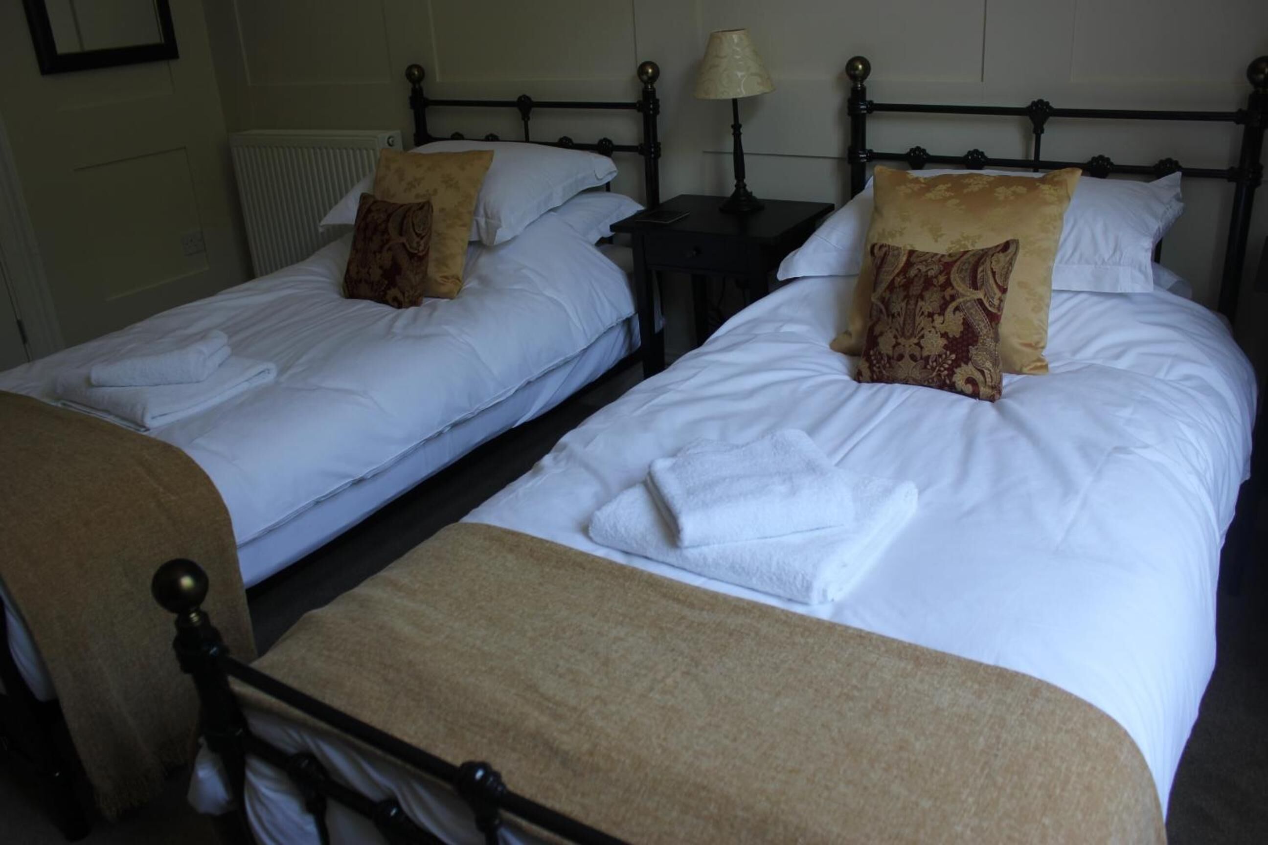 Un lit double avec du linge de lit blanc et deux coussins décoratifs, une table de chevet avec une lampe et un radiateur au mur.