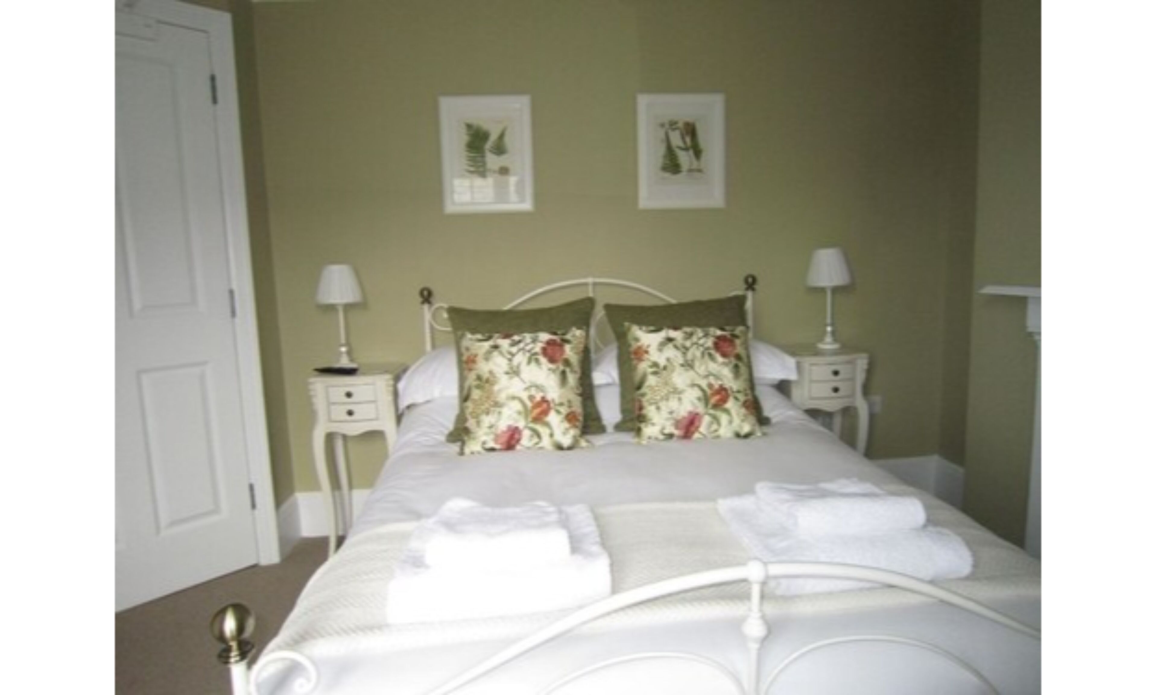 Una cama bien hecha con almohadones florales y dos mesitas de noche con lámparas, en una habitación con una pared lisa y una puerta.