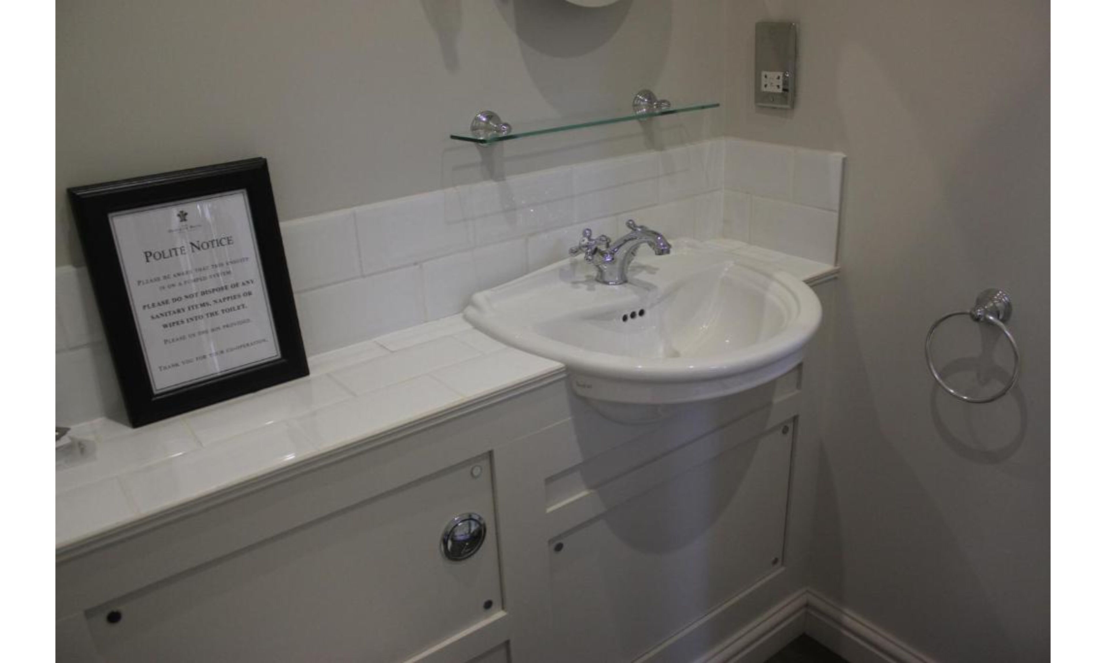 A bathroom with a white sink, a framed notice, and a glass shelf.
