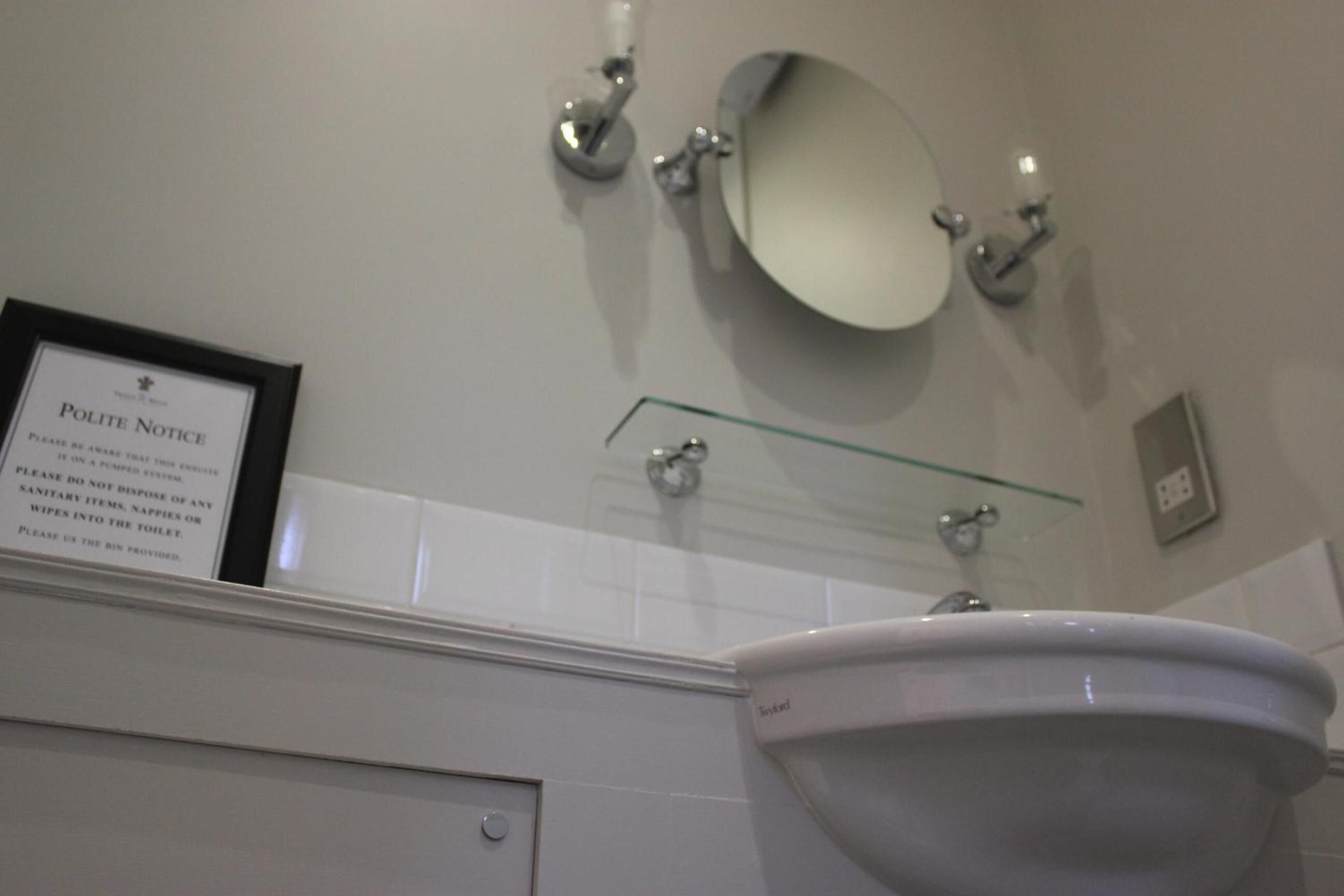 A bathroom with a white sink, a framed polite notice, a glass shelf, and a mirror.