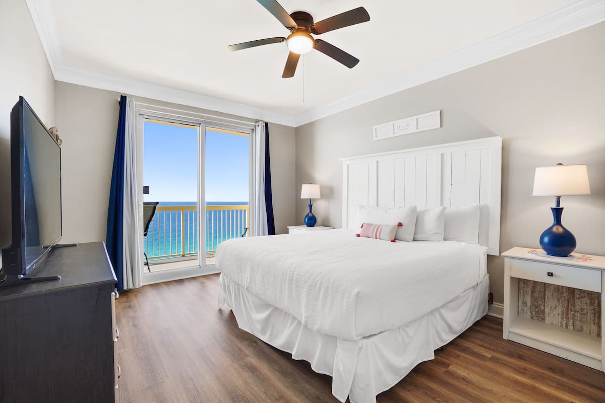 A bedroom with a large bed, a ceiling fan, a TV, and a view of the ocean through a sliding door.