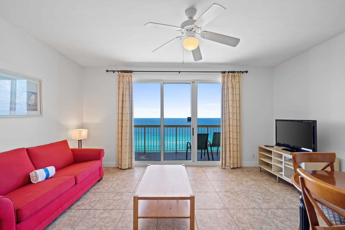 A room with a red sofa, a wooden table, a TV on a stand, and a balcony with a view of the ocean.