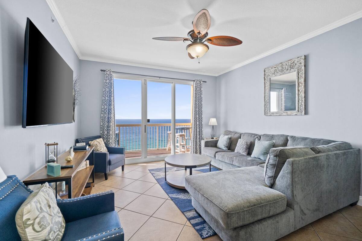 A spacious living room with a large grey sofa, a blue armchair, and a coffee table. A ceiling fan is visible, and there is a large window offering a view of the ocean.