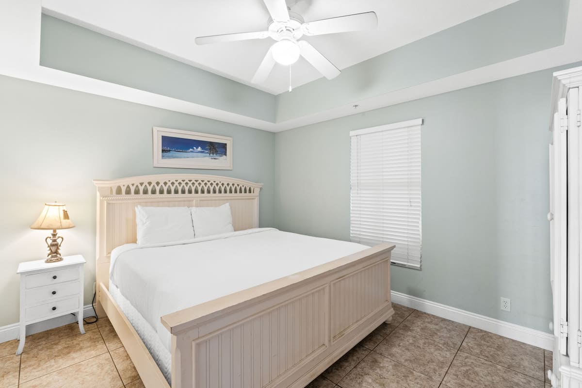 A bedroom with a bed, a nightstand, a ceiling fan, a window with blinds, and a door.
