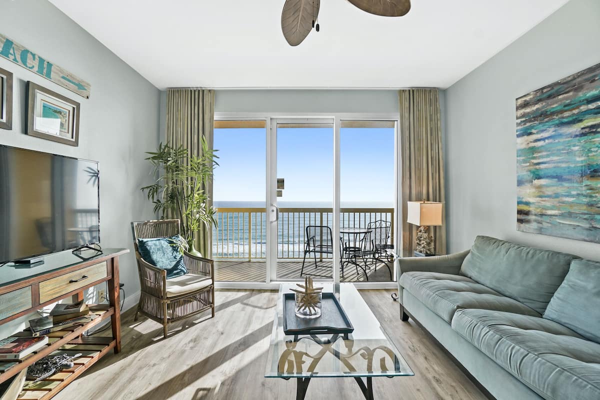 A living room with a sofa, a coffee table, a TV, and a balcony with a view of the ocean.