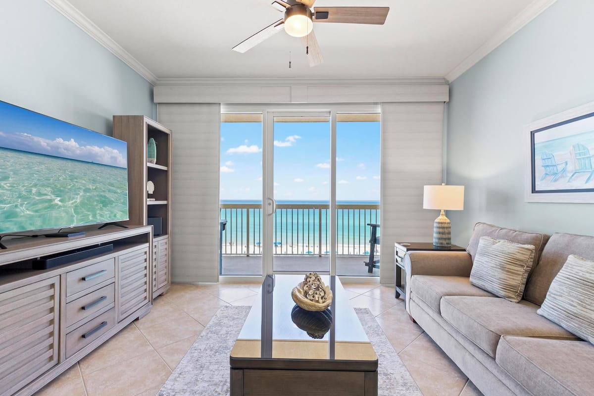 A living room with a TV, a coffee table, a sofa, a ceiling fan, and a balcony with a view of the beach.