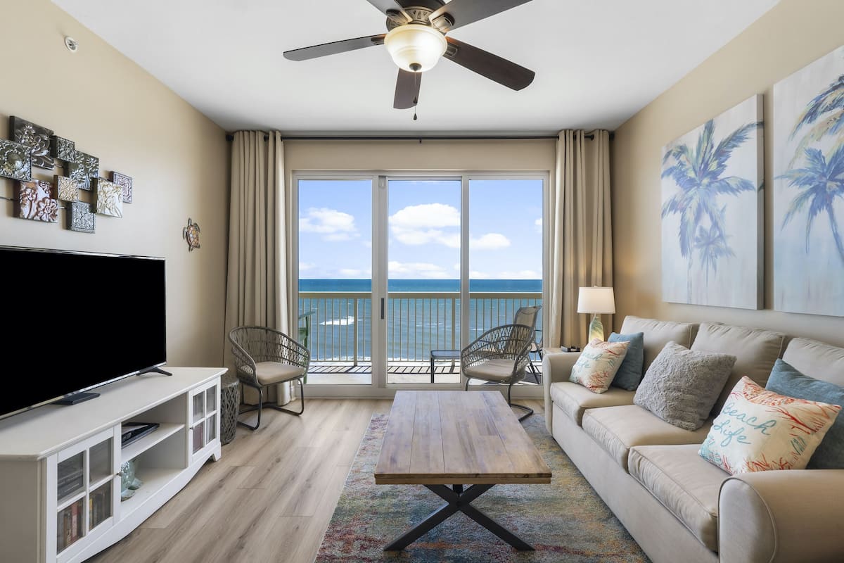 A living room with a TV, a ceiling fan, a sofa, and a view of the ocean.