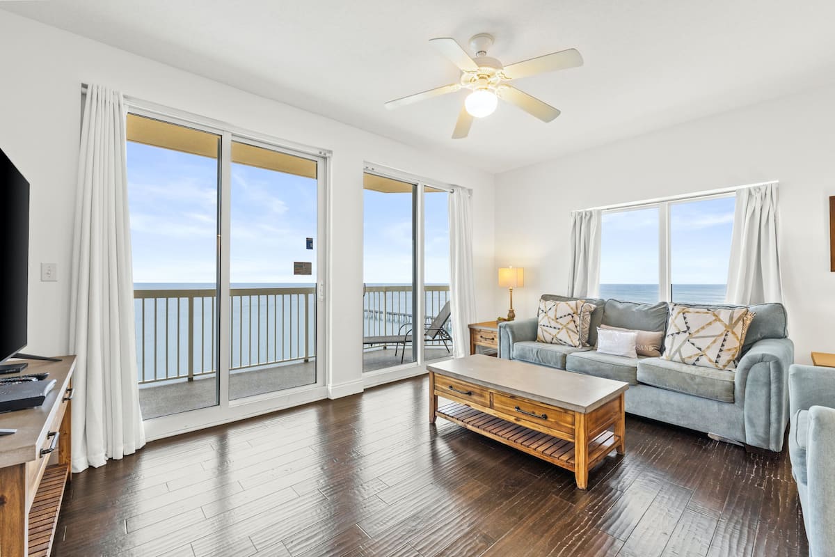 A spacious living room with a large sliding glass door leading to a balcony, a ceiling fan, a wooden coffee table, a grey sofa with patterned cushions, and a TV on a wooden stand.