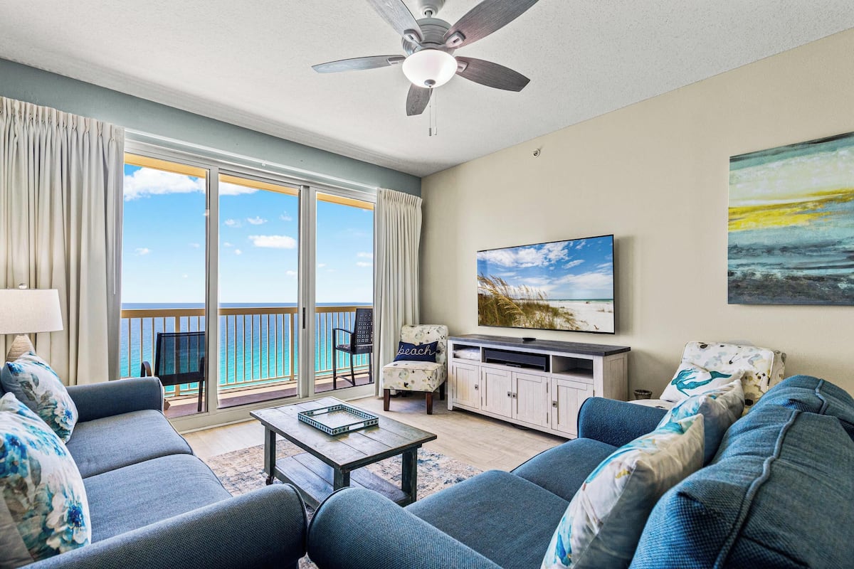 A living room with a sofa, coffee table, TV, and a balcony with a view of the ocean.