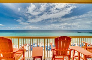 Ein Balkon mit Holzstühlen und einem Tisch mit Blick auf einen Strand mit Sonnenschirmen und einem Steg.