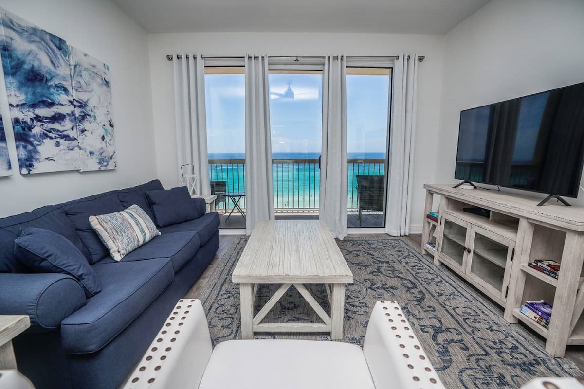 A living room with a blue sofa, a white coffee table, a TV on a wooden stand, and a view of the ocean through large windows.