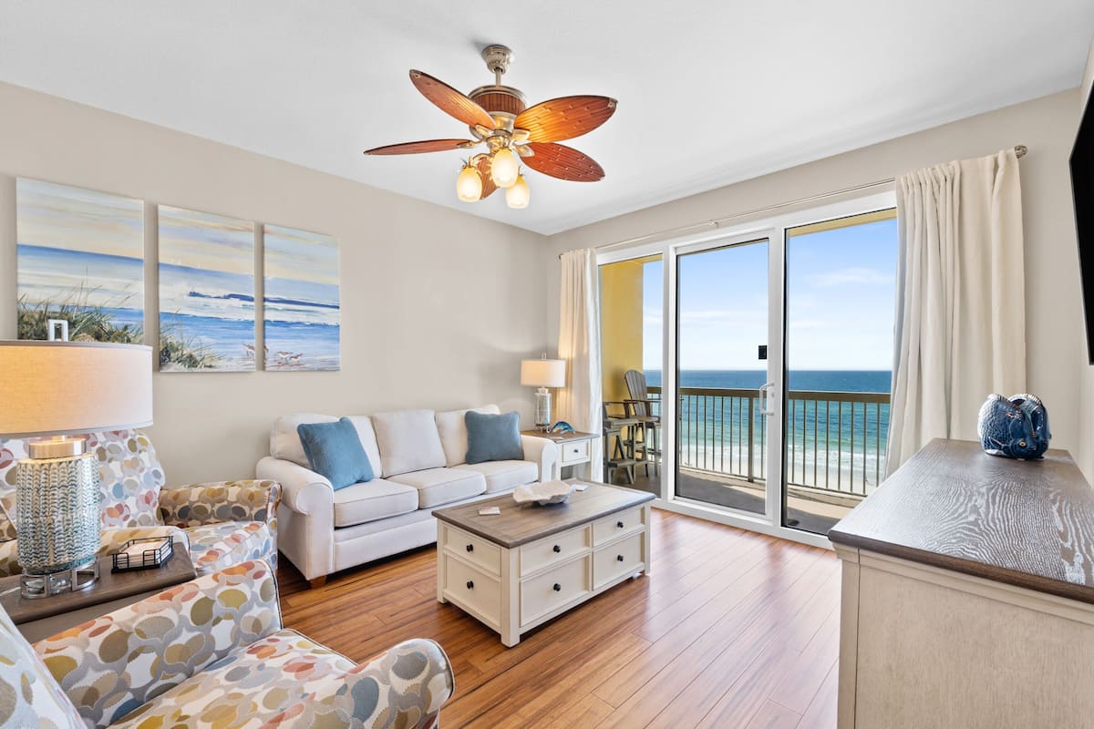 A spacious living room with a large window offering a view of the ocean, a ceiling fan, and a wooden floor.