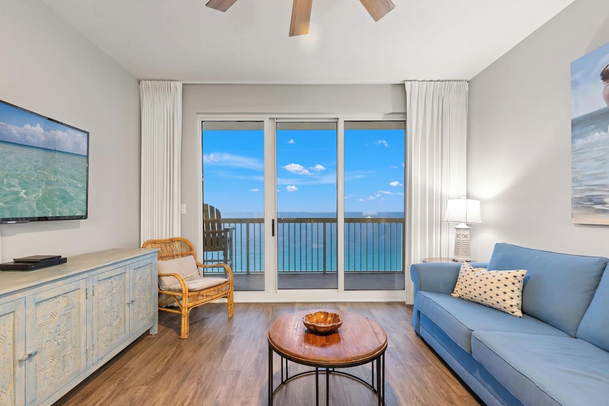 A spacious living room with a large window offering a view of the ocean, a blue sofa, a wicker chair, a small round table, and a television on a cabinet.