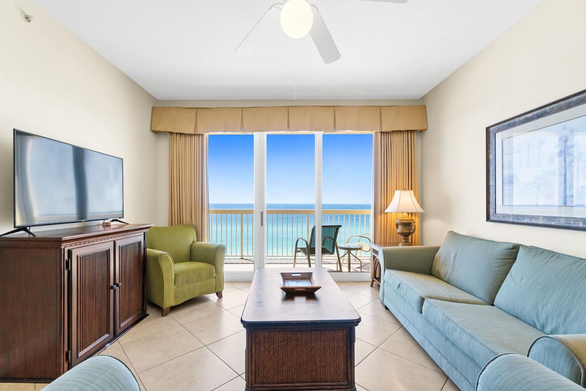 A hotel room with a large TV, a wooden cabinet, a green armchair, a blue sofa, a small table, and a balcony with a view of the ocean.