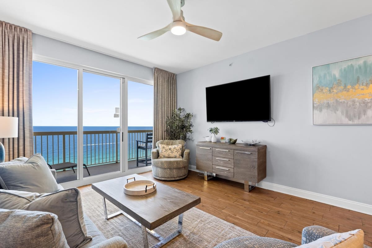 A modern living room with a balcony overlooking the sea, a flat-screen TV, and a dining area.
