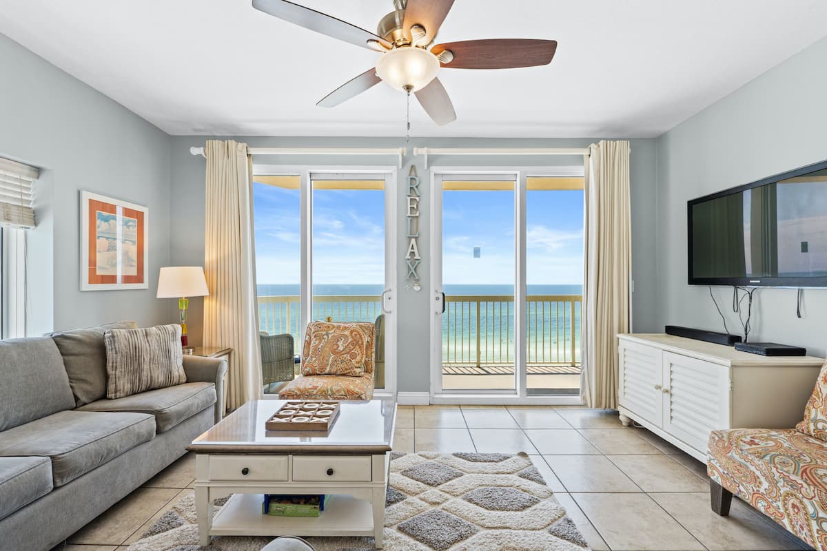 A living room with a sofa, armchair, coffee table, TV, and a balcony with a view of the ocean.