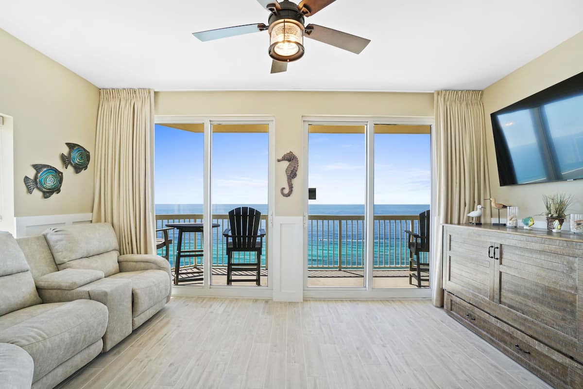 A spacious living room with a large sofa, a ceiling fan, and a view of the ocean through sliding glass doors.