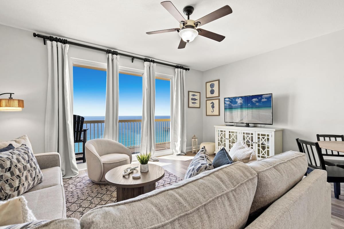 A living room with a sofa, armchairs, a coffee table, a TV, and a balcony with curtains.